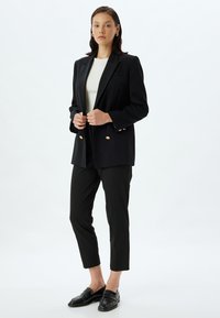 Zwarte op maat gemaakte blazer met gouden knopen, gecombineerd met strakke zwarte broek en glanzende loafers. Witte top eronder; strak ontwerp.