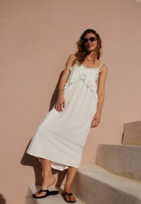 Robe maxi blanche sans manches avec des volants sur le buste. Texture lisse. Portée avec des tongs noires, debout sur des marches contre un mur rose.