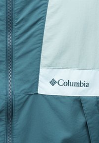 Κοντινό πλάνο ύφασματος μπουφάν σε τόνους τιρκουάζ και ανοιχτού γκρι με εμφανές το λογότυπο Columbia και το φερμουάρ στο εμπρός μέρος.