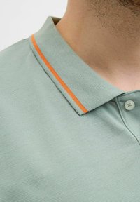 Hele roheline polo särk tekstureeritud pinnaga, millel on kaelus oranžiga aktsentiribaga ja plastiknuppude kinnitamine.