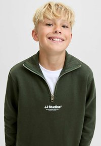 Grön zip-up sweatshirt med hög krage, med en vit logotyp på bröstet. Slät textur, avslappnad passform, lager över en vit skjorta.