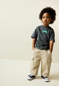 Szürke rövid ujjú póló "COOL" grafikával, bézs cargo nadrággal és fekete, fehér talpú sportcipőkkel párosítva.