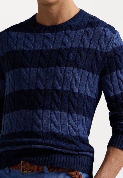 Marineblå kabelstrikket sweater med vekslende mørkeblå og lyseblå striber, ribbestrikket hals og manchetter, samt et lille emblem på brystet.