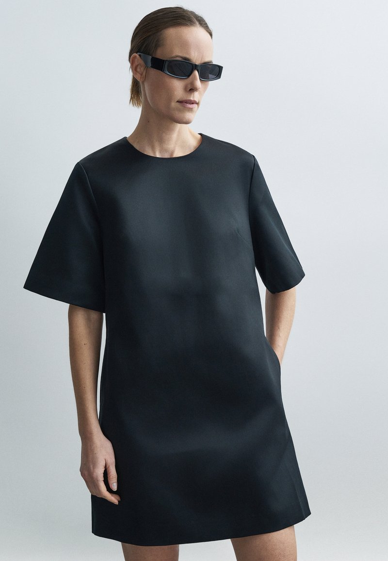 Robe noire à manches courtes en tissu lisse, dotée d'un col rond et de poches latérales. Le modèle porte des lunettes de soleil fines noires.