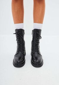 Botas de combate de cuero negro con un acabado suave, puntera redonda, cordones en la parte delantera y cremalleras en los laterales para facilitar su uso. Calcetines blancos hasta el tobillo visibles.