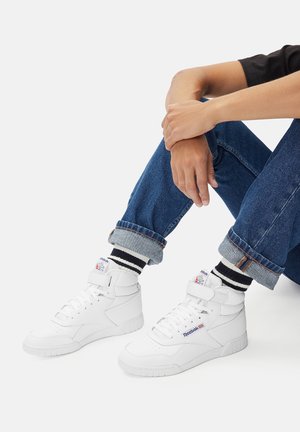 Białe sneakersy typu high-top z teksturowaną skórzaną powierzchnią, wyposażone w pasek na rzep, wyściełany kołnierz oraz logo. Noszone z podwiniętymi dżinsami.