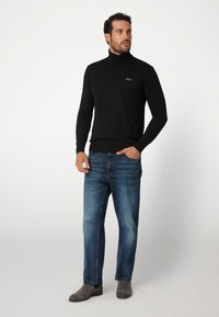 Maglione nero a collo alto con maniche lunghe, abbinato a jeans in denim blu. L'outfit è completato da stivali alla caviglia grigi e un logo del marchio discreto.