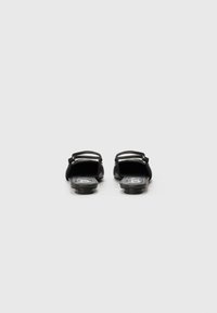 Sandale negre plate cu spatele deschis, realizate dintr-un material din piele netedă și având un design minimalist. Un mic logo vizibil pe branț.