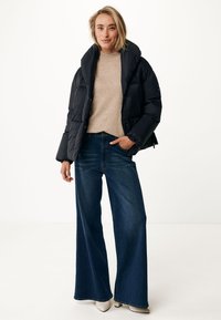 Doudoune noire avec un col haut, pull beige et jean bleu à jambes larges. La tenue comprend des bottines à talons crème. Fond uni.