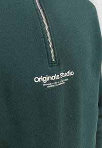 Tmavě zelená mikina se stříbrným zipem do poloviny a bílým textem "Originals Studio" a menším pruhem pod ním na oblasti hrudníku.
