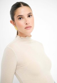 Top beige a collo alto con colletto arricciato, texture liscia, maniche lunghe e design aderente. Orecchini geometrici dorati aggiungono un dettaglio notevole.