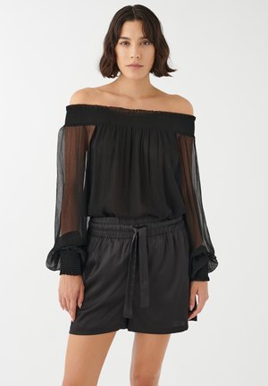 Juodas off-the-shoulder topas su permatomis ilgomis rankovėmis ir smulkinta iškirpte, derinamas su juodais šortais su surišamu juosmeniu.