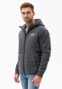Chaqueta acolchada gris con capucha, que presenta una cremallera frontal, dos bolsillos laterales y un parche de logo. La tela texturizada contrasta con las suaves mangas de forro polar.