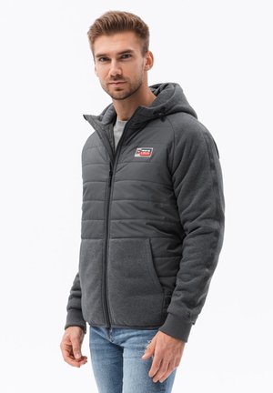 Veste matelassée grise avec capuche, dotée d'une fermeture éclair à l'avant, de deux poches latérales et d'un patch logo. Le tissu texturé contraste avec les manches en polaire douce.
