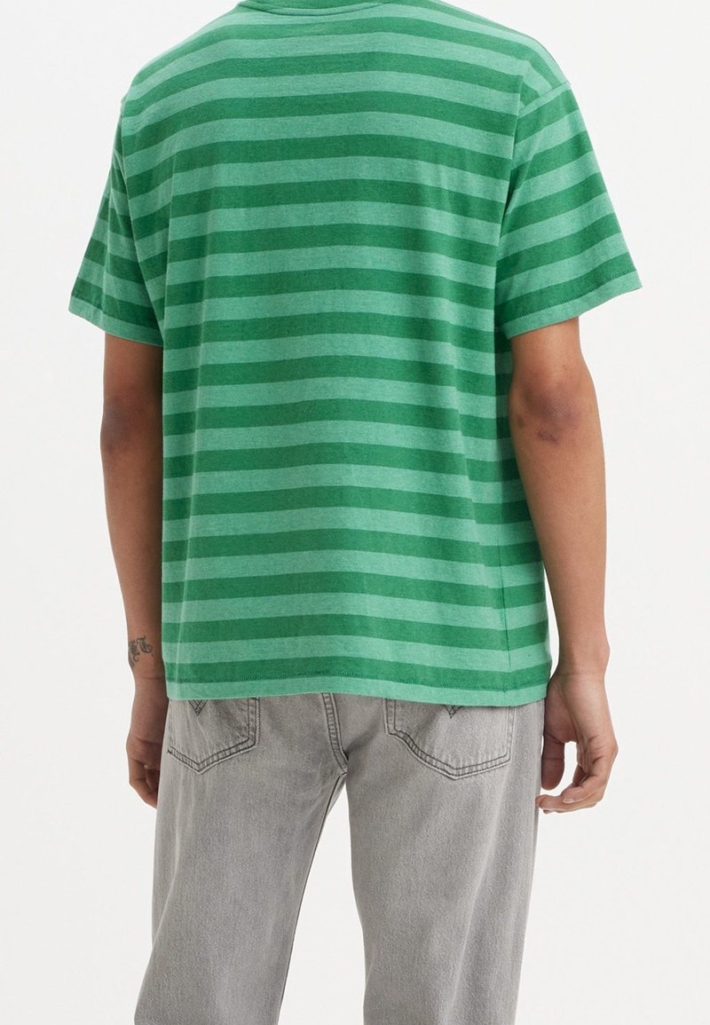 Personne portant une chemise à manches courtes rayée verte et un jean gris clair, vue de dos sur un fond blanc.