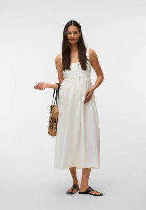 Femme en robe midi blanche sans manches tenant un sac cabas tissé, portant des sandales noires à enfiler, debout devant un fond uni.