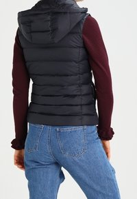 Svart pufferväst med huva, med horisontella kviltade sektioner och en figursydd design, bärs över en bordeaux långärmad topp och blå denimjeans.