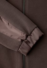 Sleeve of a brown jacket with elastic cuff and shiny fabric stripe, next to a partially visible metal zipper on the garment front.