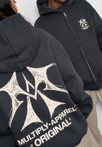 Felpa nera con zip realizzata in un mix di cotone. Presenta una grande grafica bianca sul retro con un logo stilizzato e testo in un carattere audace.