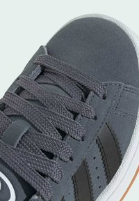 Zapatillas de ante gris con un upper texturizado, cordones planos de tejido, rayas negras y una suela de goma blanca con acentos sutiles.