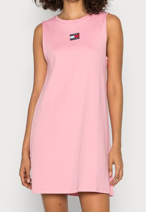 Mouwloze roze casual jurk met een klein Tommy Jeans-logo geborduurd in het midden op de borst, gedragen door een staand persoon.