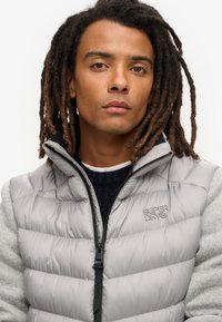 Gilet rembourré gris avec fermeture éclair et col haut, avec un tissu tricoté texturé gris clair sur les côtés. Logo de la marque sur la poitrine.
