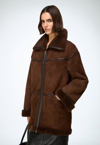 Chaqueta de ante marrón con forro de borrego, con un gran cuello, cierre de cremallera y bolsillos laterales; cuenta con detalles de cuero y un acabado suave y texturizado.