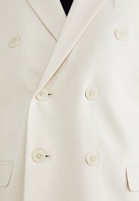 Blazer de doble botonadura en un tejido beige claro, con textura suave, seis botones a juego y dos bolsillos frontales.