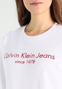 Felpa bianca in cotone misto, con testo ricamato in rosso "Calvin Klein Jeans dal 1978" sul davanti, colletto a girocollo classico.