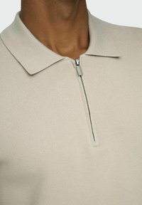 Beige Strickpolo mit Reißverschluss vorn, strukturiertem Kragen und einer strukturierten Stoffoberfläche. Es zeichnet sich durch einen subtilen Glanz und saubere Verarbeitungsdetails aus.