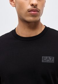 Mladi moški, ki nosi črno T-majico z okroglim izrezom in majhnim logotipom EA7 Emporio Armani na levi strani prsi, posneto od brade do ramen.