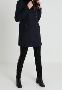 Person som bär en mörk kappa med pälskrage, svarta byxor med sidostråk och svarta snörade ankelstövlar, stående mot en slät bakgrund.