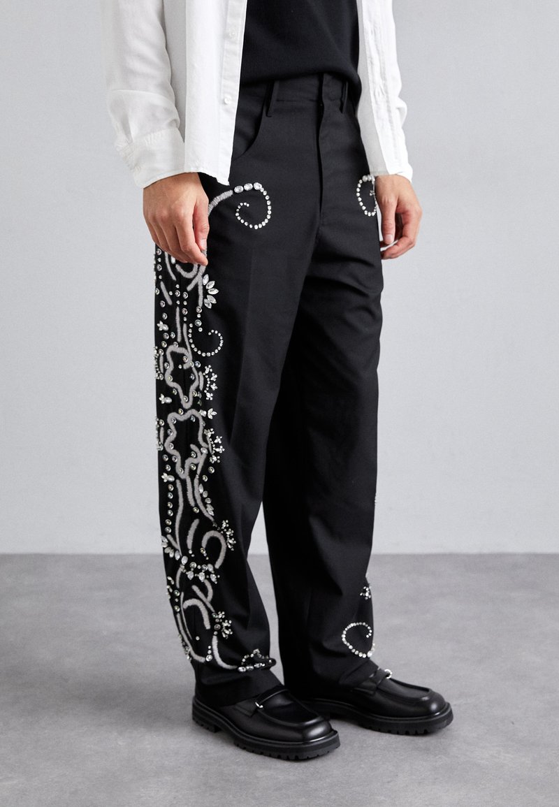 Fekete nadrág, amelyet bonyolult ezüst hímzés díszít, hullámzó mintákkal és dekoratív elemekkel az oldalán, fekete cipővel párosítva.