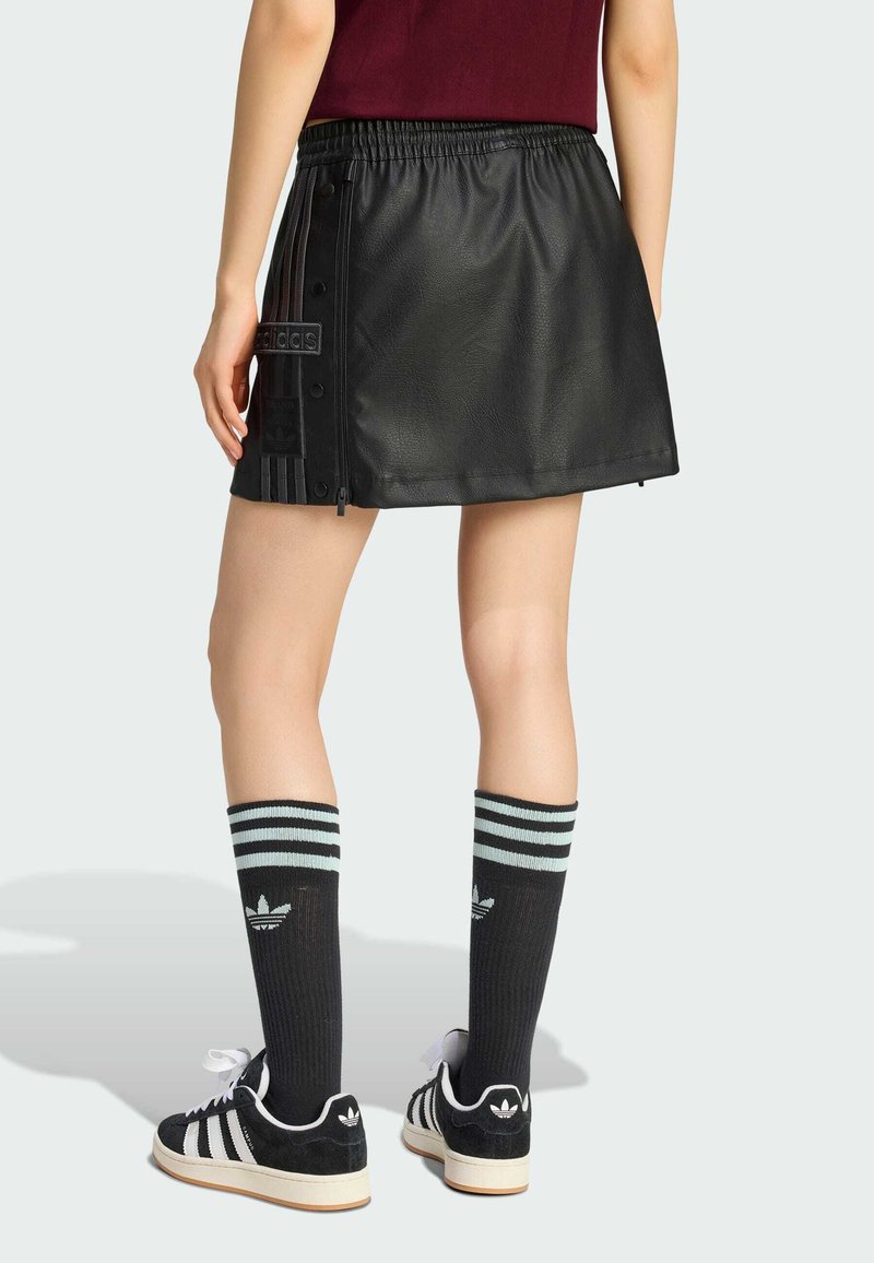 Person wearing a black leather mini skirt with side snap buttons, black Adidas crew socks with stripes, and black Adidas sneakers.