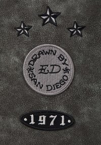 Gray textured fabric features three embroidered stars above a circular patch stating “DRAWN BY ED SAN DIEGO” and a lower oval badge with “1971.”