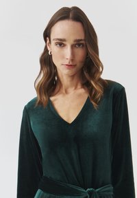 Dark green velvet top with long sleeves and a V-neckline, featuring a belted waist detail and a soft, textured finish.