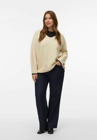 Femme portant un pull oversized crème, un jean large noir et des chaussures noires, se tenant avec une main dans la poche sur fond blanc.