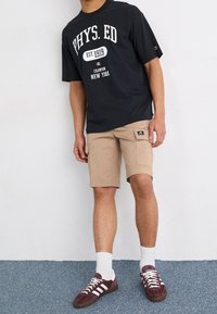 Muž v černém tričku s nápisem "FYZ. VZD" a béžových cargo šortkách, bílých ponožkách a vínových teniskách Adidas stojí na modrém koberci.