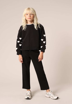 Sweatshirt noir avec motifs de cœurs blancs sur les manches, associé à un pantalon noir et des baskets gris clair. Tissu lisse, coupe décontractée.