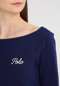 Žena ve tmavě modrém topu se širokým výstřihem a bílým vyšívaným textem "Polo" na hrudi, na jednolitém světlém pozadí.