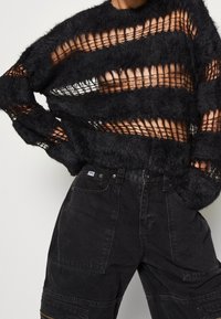 Black fuzzy knit sweater with open stripes over a black cargo pant featuring multiple pockets and a tag on the waistband.