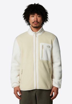 Fleece jacket in cream and white, featuring a high collar, front zipper, and a zippered chest pocket with a logo. Soft texture and casual fit.