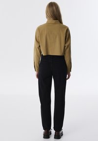 Camisa corta de color verde oliva con yugo trasero, combinada con jeans negros de cintura alta y complementada con zapatos marrones con cordones. Diseño minimalista.