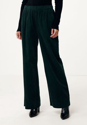 Pantalon large en velours côtelé vert foncé porté avec des bottines à talons noires et un haut noir à manches longues, debout sur fond blanc.