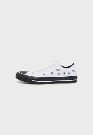 Zapatilla blanca de corte bajo con suela y puntera negras, decorada con pequeñas tachuelas metálicas plateadas en forma de murciélago, gato y lazo en los laterales.
