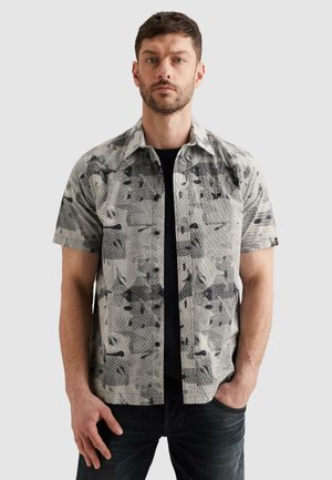 Mężczyzna z brodą w krótkim rękawem koszuli z szaro-czarnym abstrakcyjnym wzorem na czarnej koszulce, stojący z jedną ręką w kieszeni.