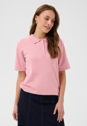 Femeie cu păr lung și castaniu, purtând cercei cu perle, un tricou polo roz cu mâneci scurte și o fustă din denim închis, privind în jos și zâmbind ușor.