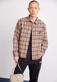 Brown plaid flannel shirt with button closure and chest pocket, paired with black pants and a beige crossbody bag. Textured fabric.