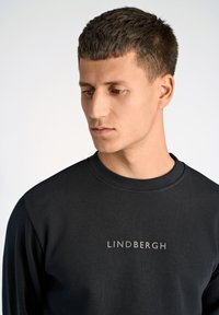 Musta collegepaita, jossa on pyöreä pääntie ja etupuolella painettu 'LINDBERGH' -logo hopeanvärisenä, sileä kangaspinta.