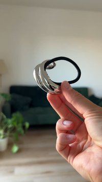 Srebrny metalowy pierścień o skręconym wzorze, z gładką, odbijającą światło powierzchnią i przymocowaną czarną elastyczną opaską. Dłoń trzymająca przedmiot.